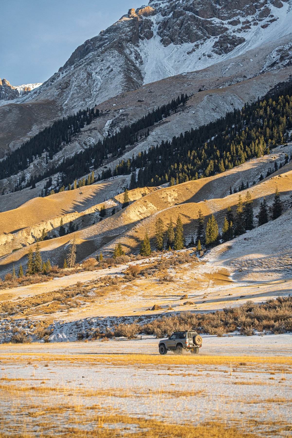 An off-road vehicle drives across a snowy field with golden grasses, surrounded by towering mountains